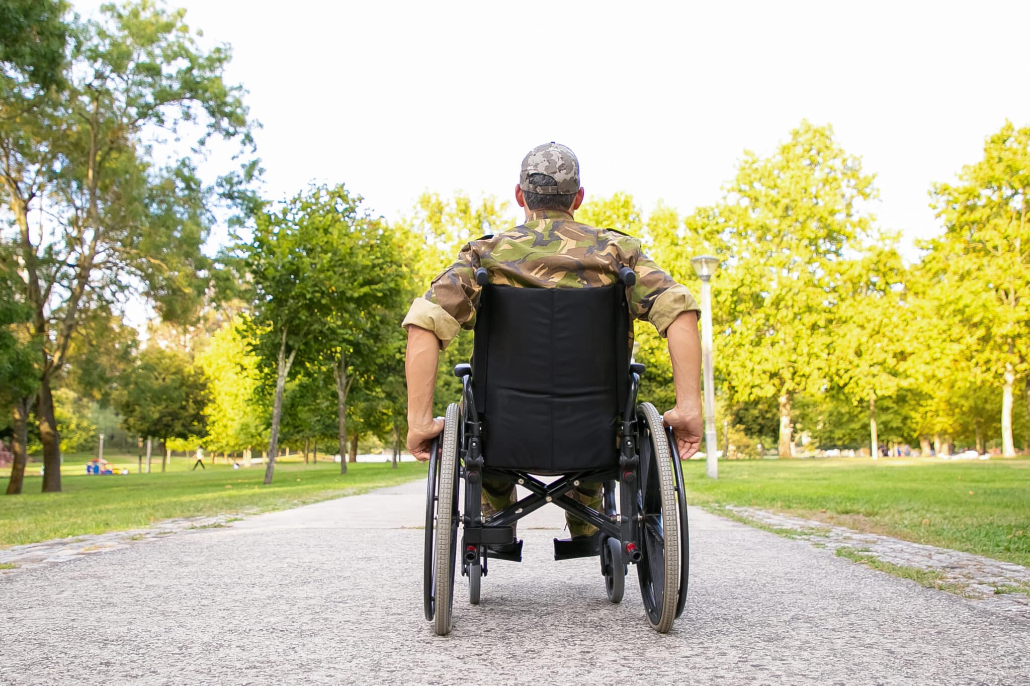 Single disabled retired military man in wheelchair moving down footpath in city park. Back view. Veteran of war or disability concept